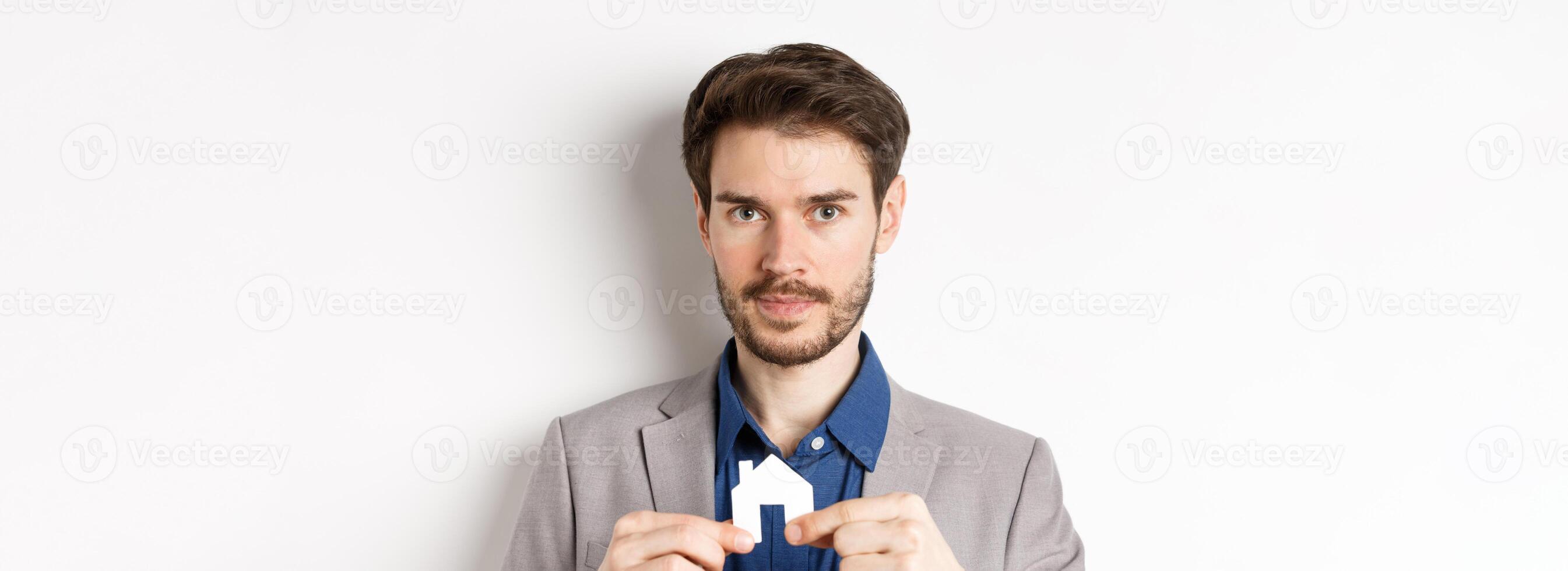 Real estate and insurance concept. Salesman realtor showing small paper house cutout and looking at camera, selling property, white background photo