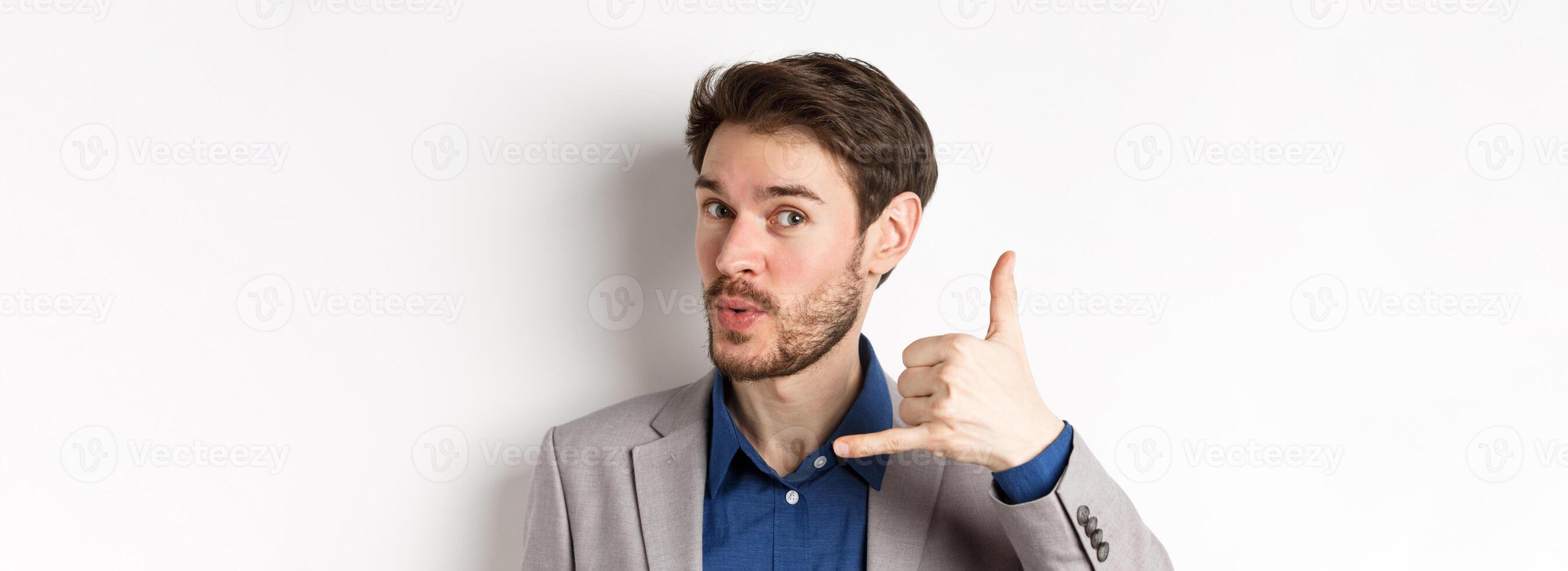 Handsome macho guy in suit showing phone sign near ear, flirting and asking to call him, giving his number, white background photo