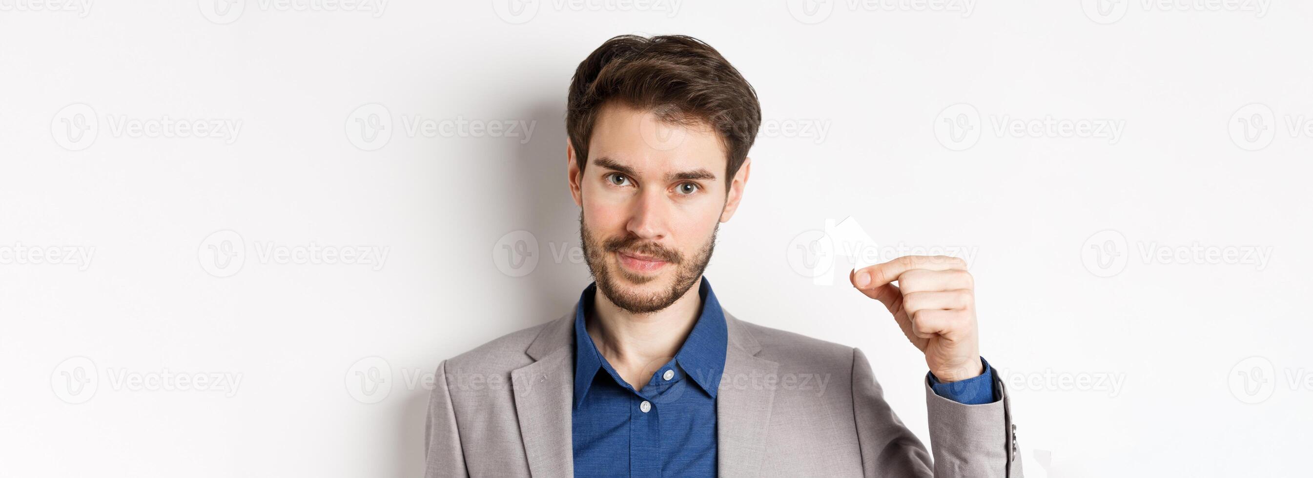 Real estate and insurance concept. Close up portrait of handsome bearded man in suit showing small paper house cutout and look assertive, white background photo