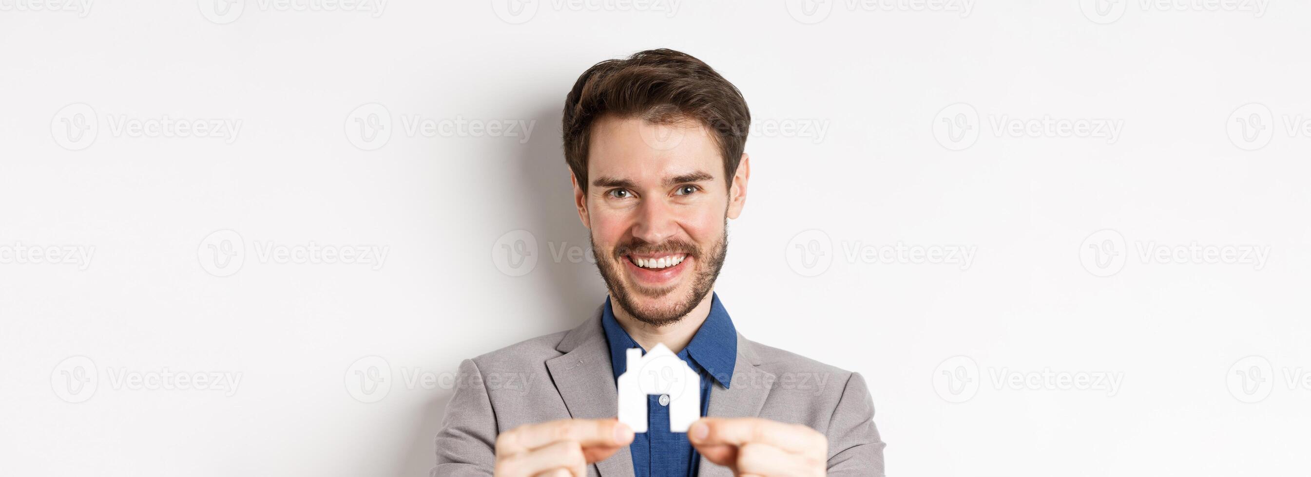 Real estate and insurance concept. Handsome man in suit smiling, showing small paper house cutout, standing on white background photo