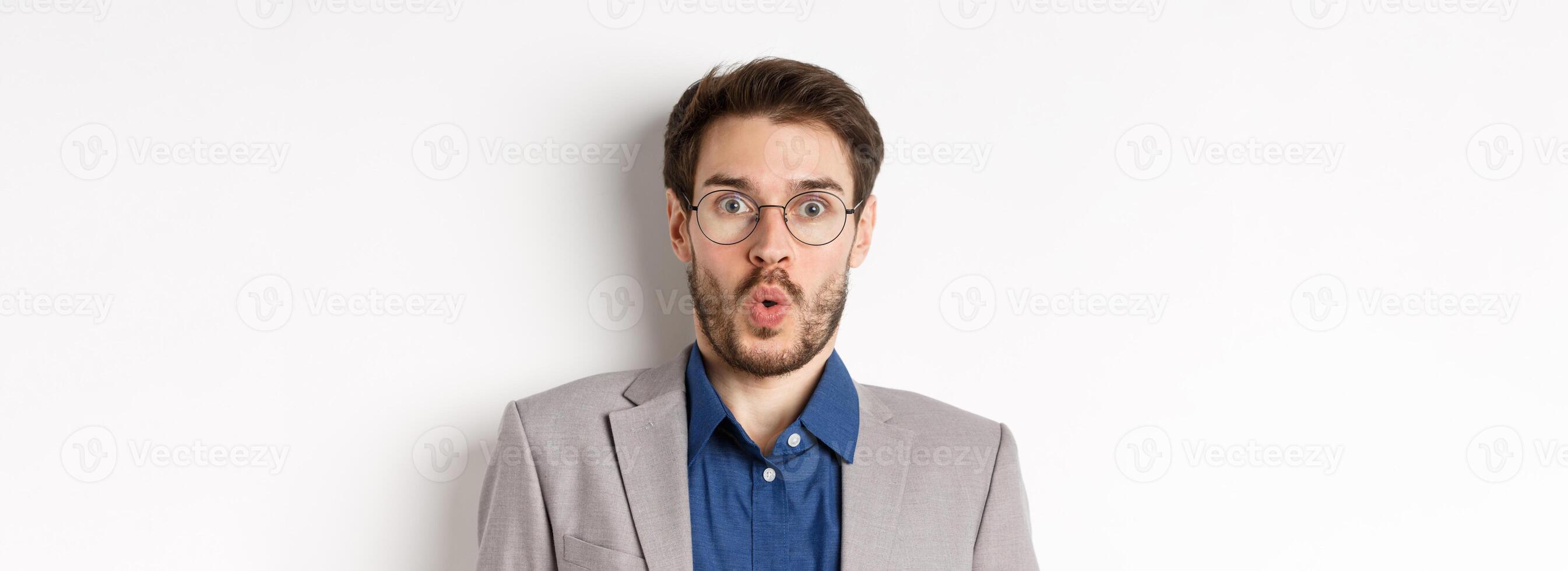 Close up portrait of excited office worker in glasses and suit saying wow, staring amazed at camera, standing against white background photo