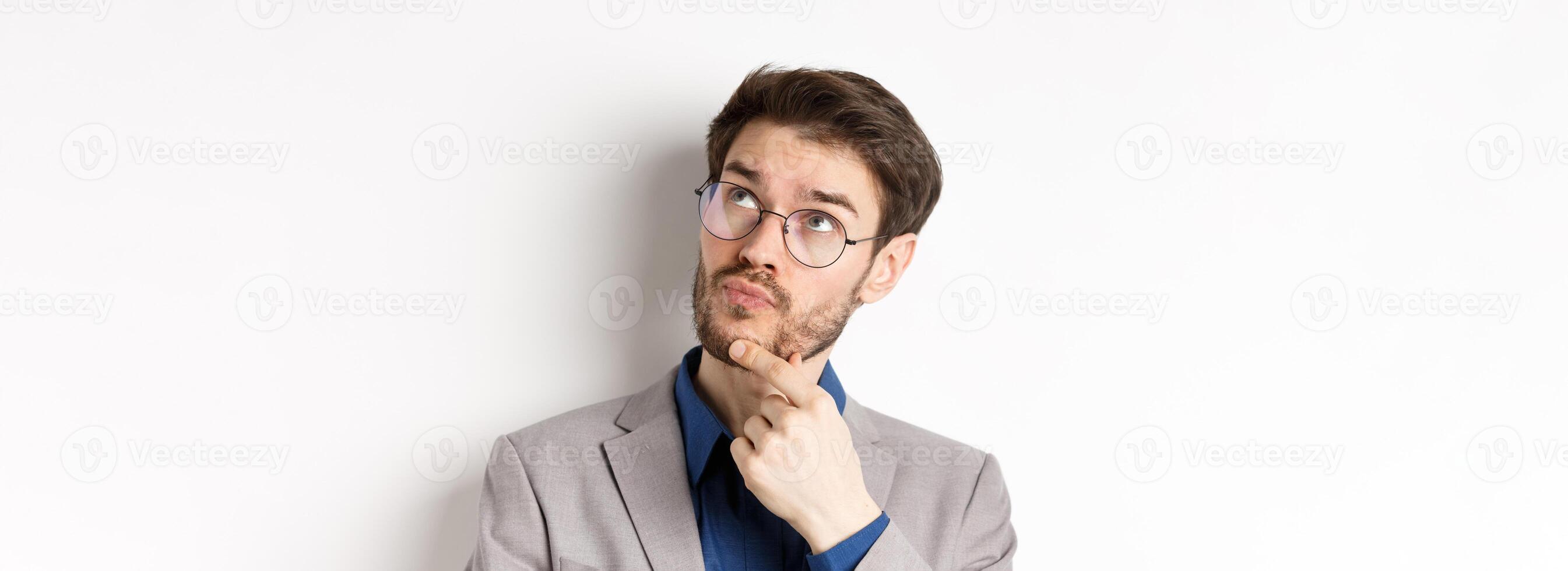 Thoughtful businessman with beard and glasses making decision, looking up and thinking, standing in suit with pensive face, white background photo