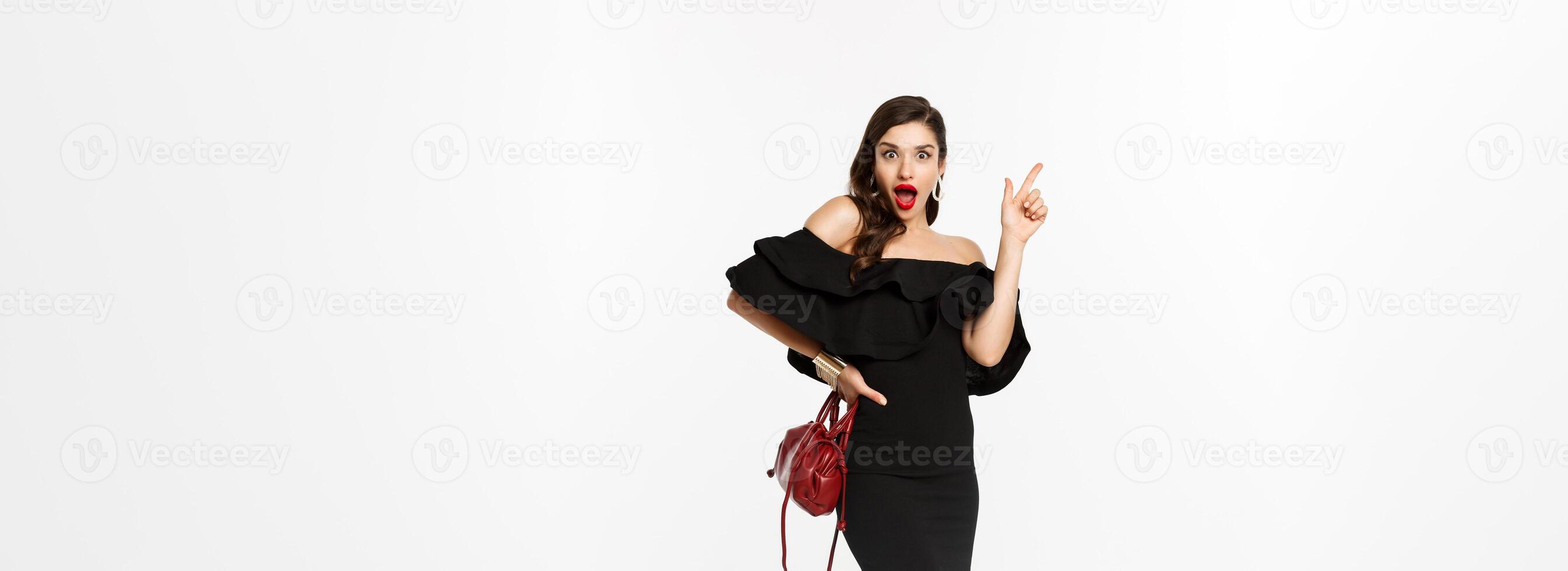 Beauty and fashion concept. Full length of excited young woman in glamour dress, red lips, having an idea, raising finger to suggest something, white background photo