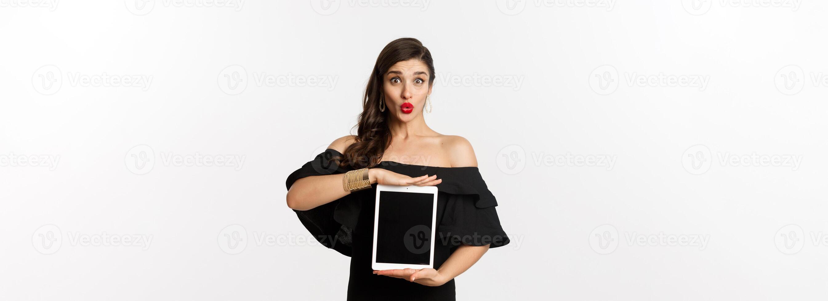 Fashion and shopping concept. Beautiful woman with red lipsticks, black dress, showing tablet screen and looking excited, standing over white background photo