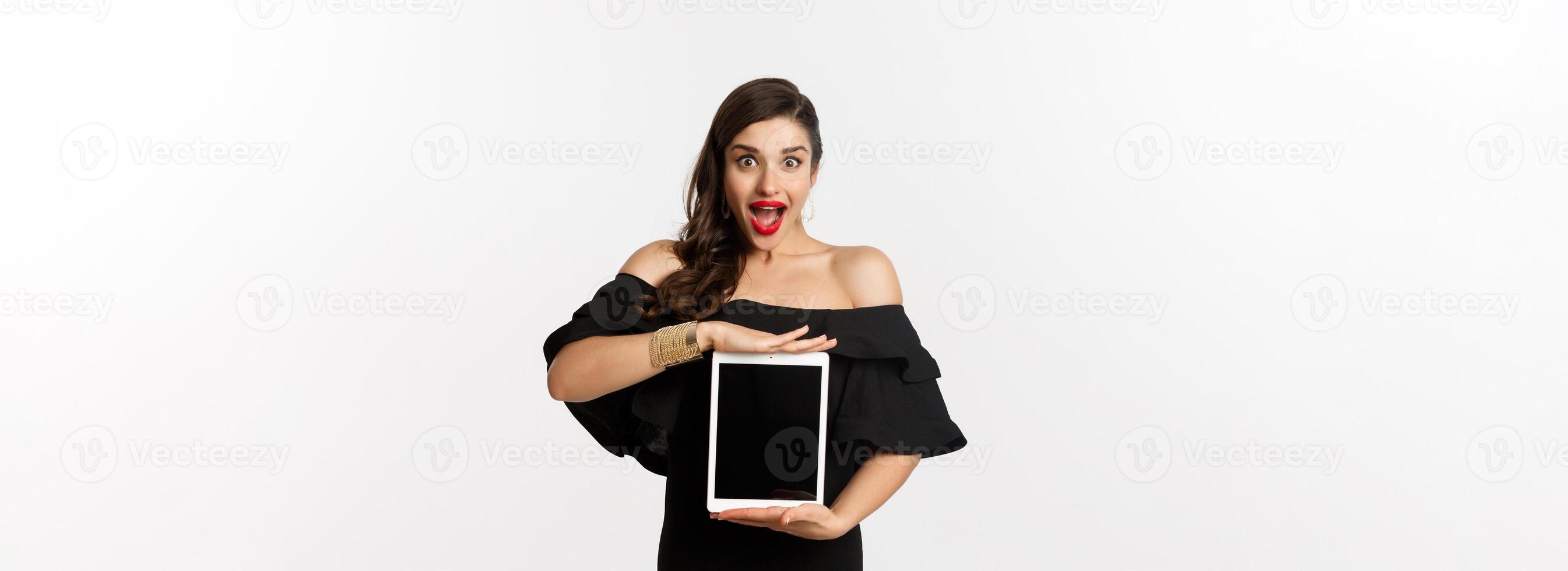 Fashion and shopping concept. Amazed young woman showing online website promo offer on tablet screen, looking excited camera, standing in black dress, white background photo