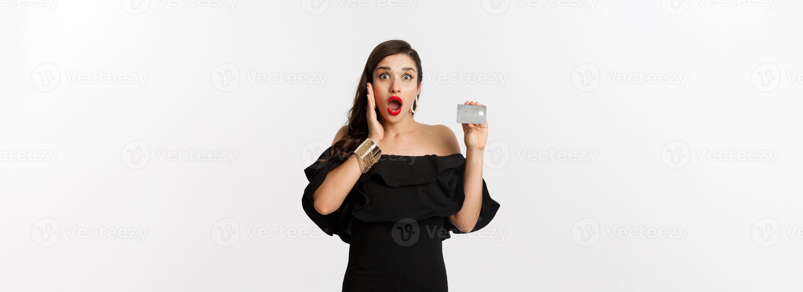 Fashion and shopping concept. Excited elegant woman with red lips, wearing black dress, showing credit card and looking amazed, standing over white background photo