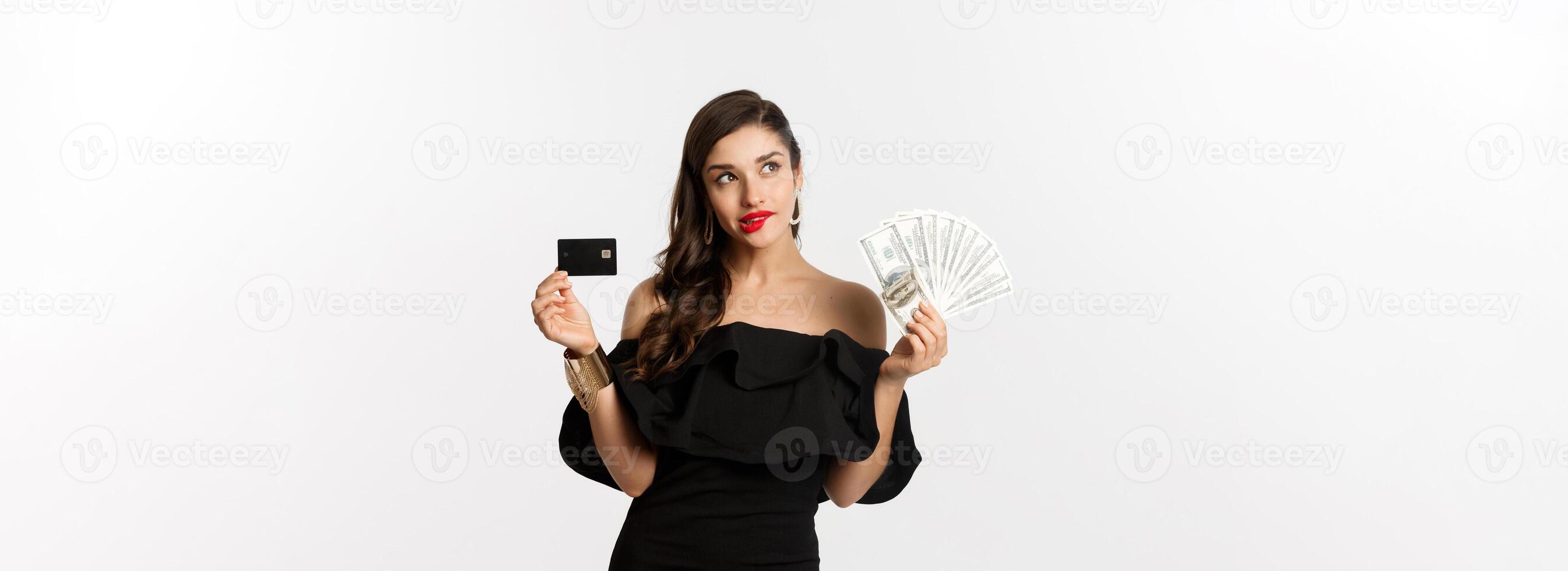 Fashion and shopping concept. Elegant woman in black dress, red lipstick, holding money and credit card, making choice, thinking, standing over white background photo
