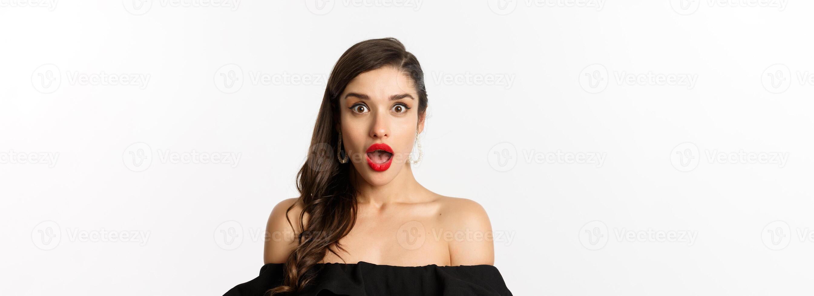 Fashion and beauty concept. Close-up of attractive brunette woman in black dress open mouth surprised, looking in awe at camera, white background photo