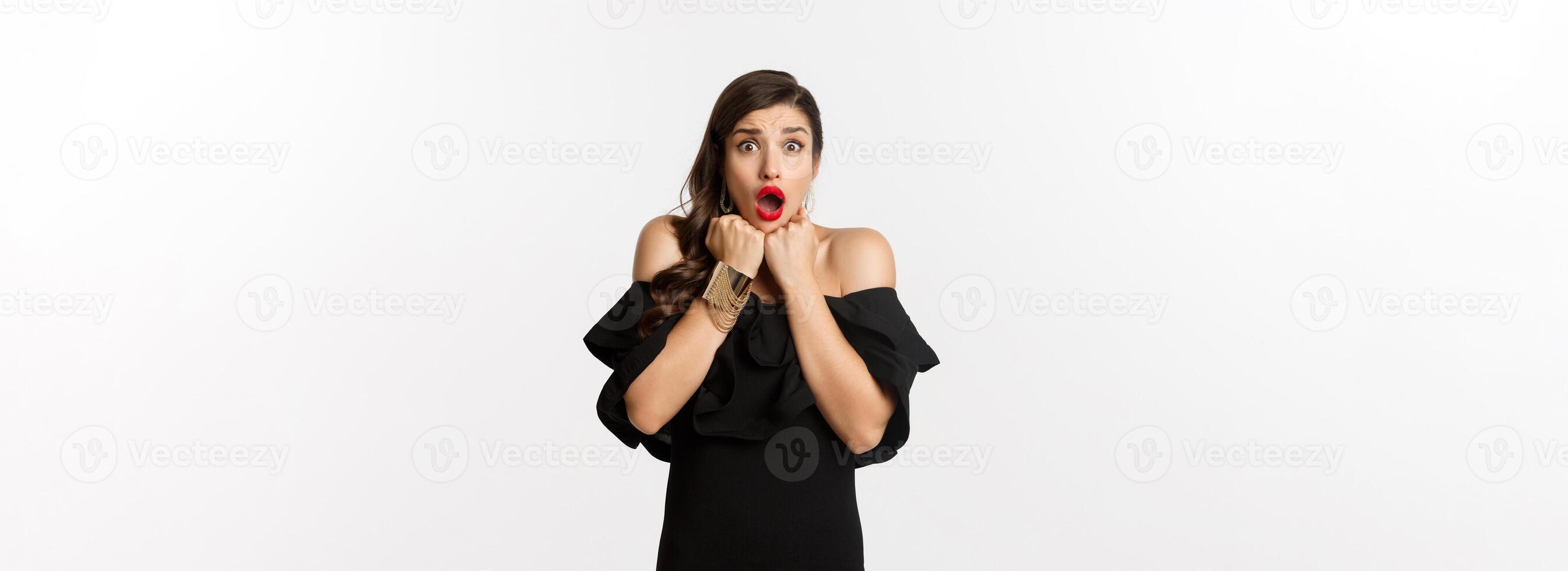 Portrait of glamour woman looking scared and shocked at camera, staring at something with fear, standing in black dress against white background photo
