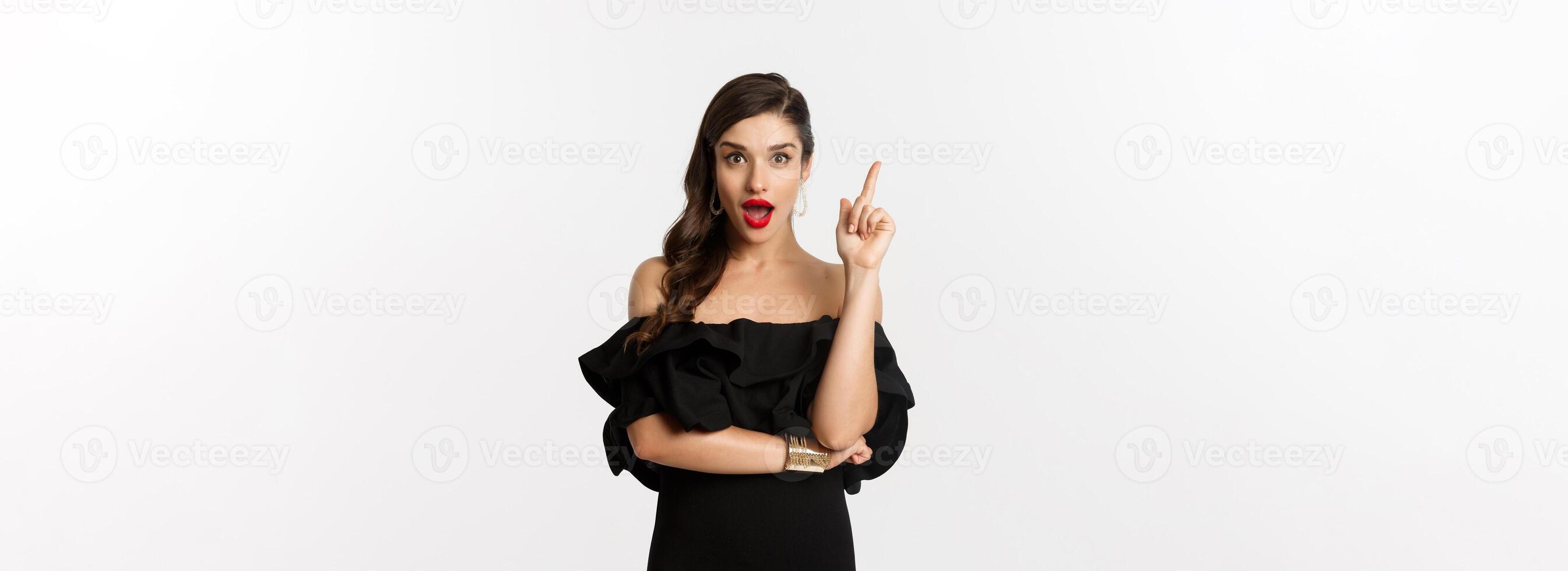 Fashion and beauty. Attractive caucasian woman in black dress having an idea, raising finger and saying suggestion, white background photo