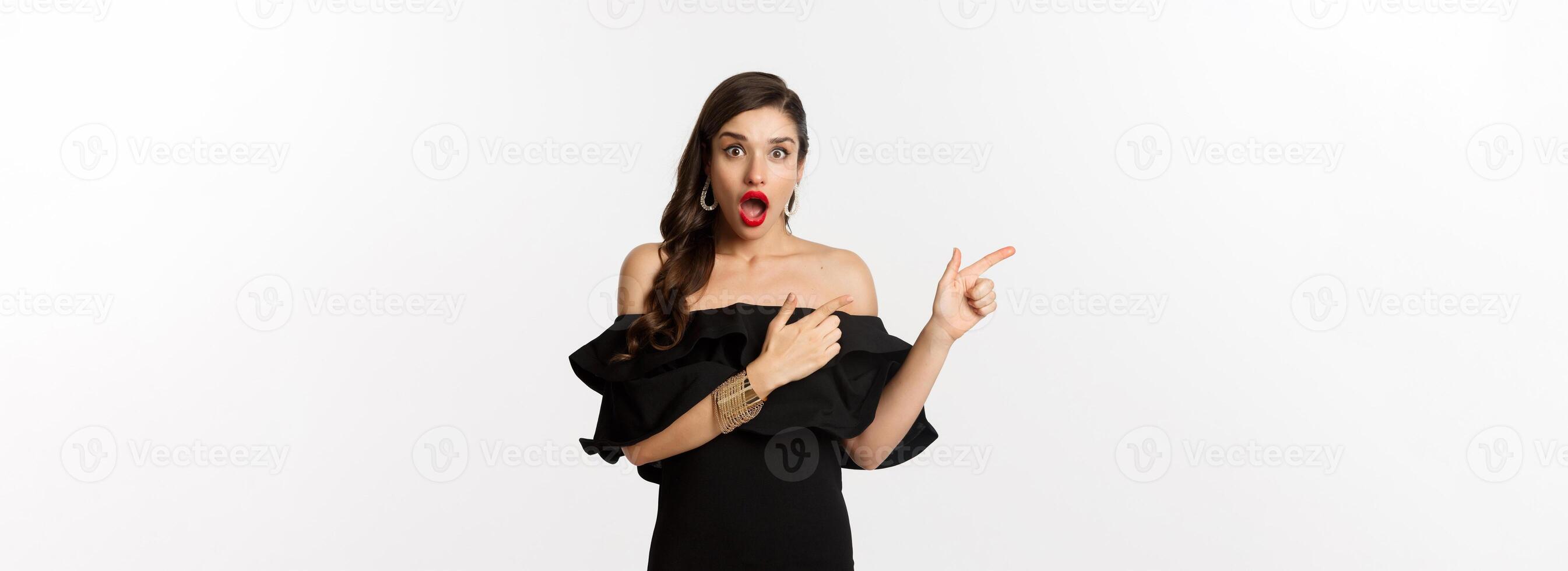 Fashion and beauty. Surprised woman in black glamour dress pointing fingers left, showing advertisement and staring amazed, white background photo