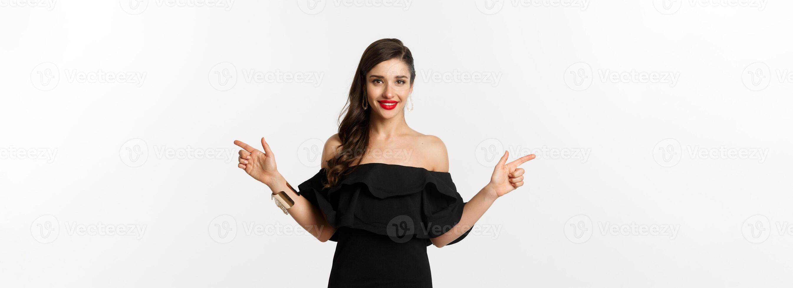 Fashion and beauty. Attractive woman in jewelry, makeup and black dress, laughing and pointing fingers sideways copy spaces offer, white background photo