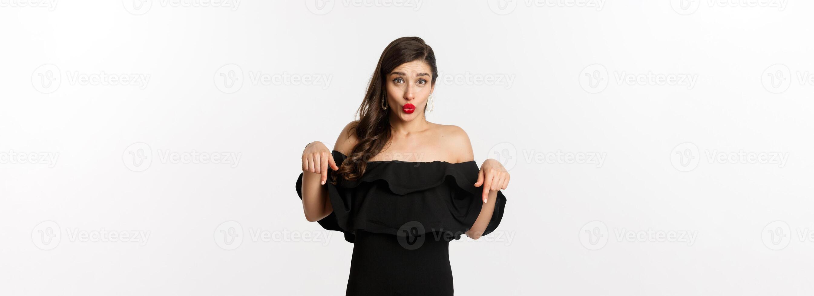 Fashion and beauty. Elegant woman in black dress pointing fingers down, showing promo and smiling, standing over white background photo