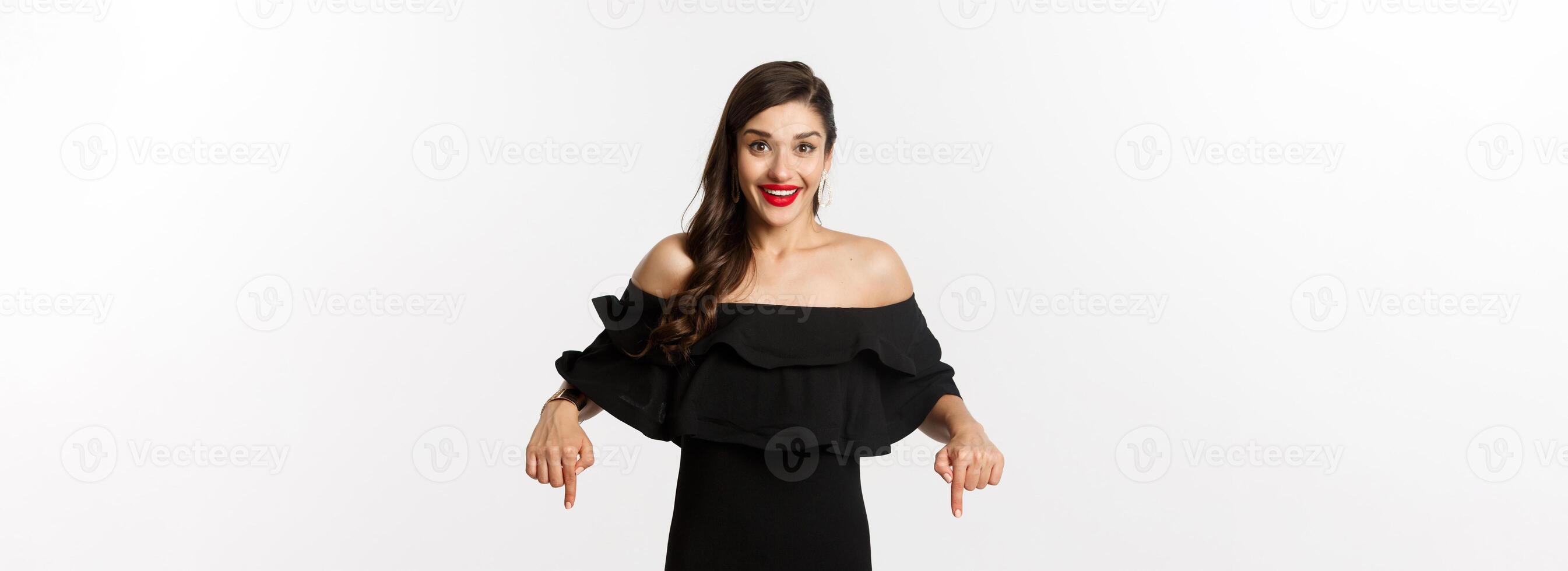 Fashion and beauty. Elegant woman in black dress pointing fingers down, showing promo and smiling, standing over white background photo