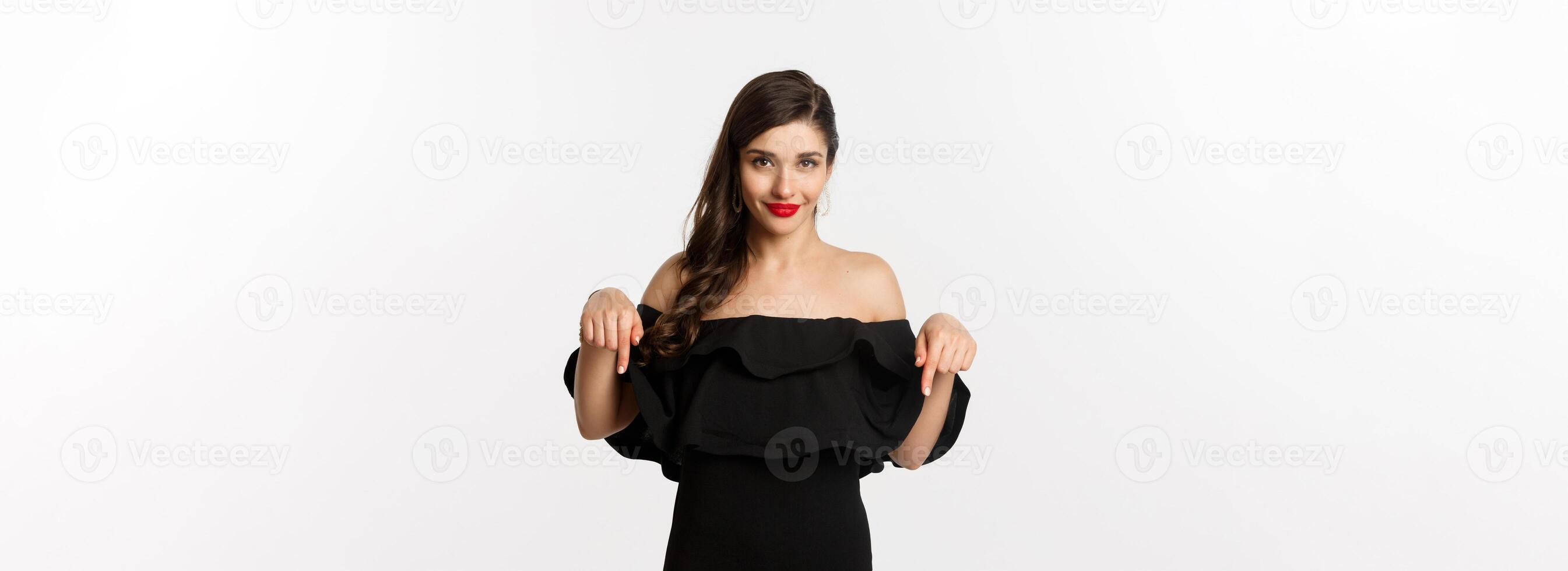 Fashion and beauty. Elegant woman in black dress pointing fingers down, showing promo and smiling, standing over white background photo