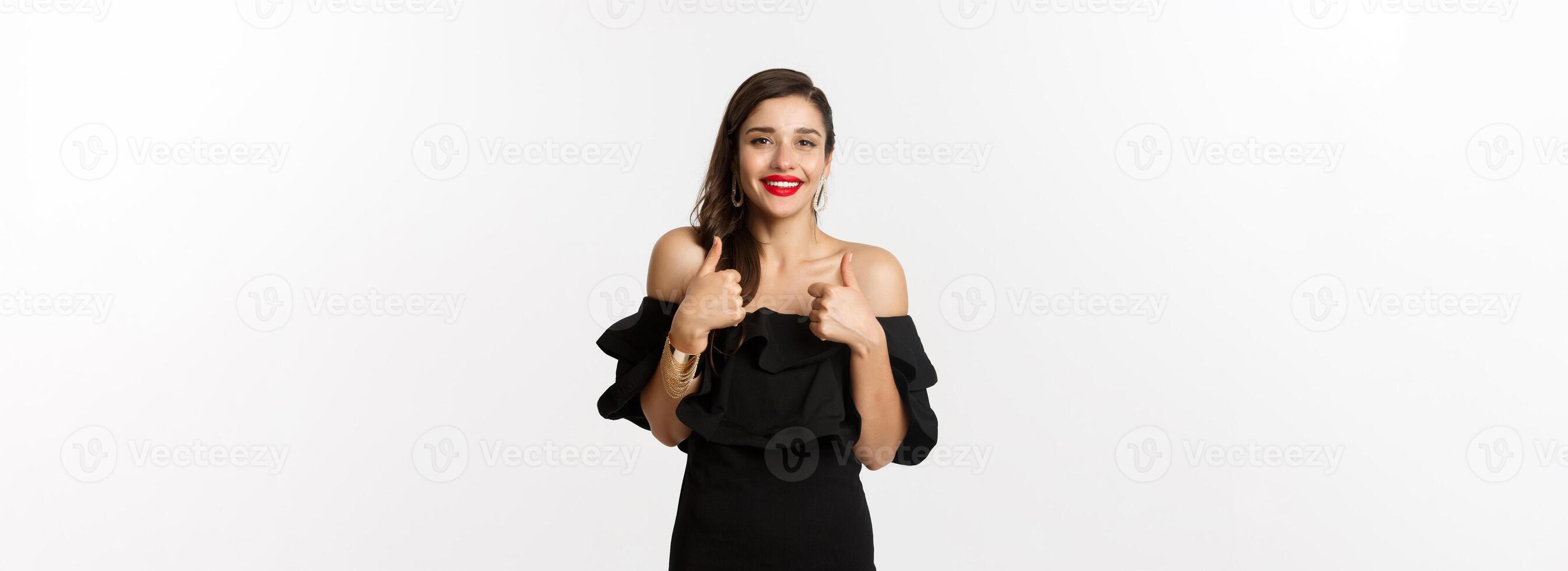 Fashion and beauty. Gorgeous woman in black dress, red lipstick, showing thumbs up in approval, recommending product, standing over white background photo