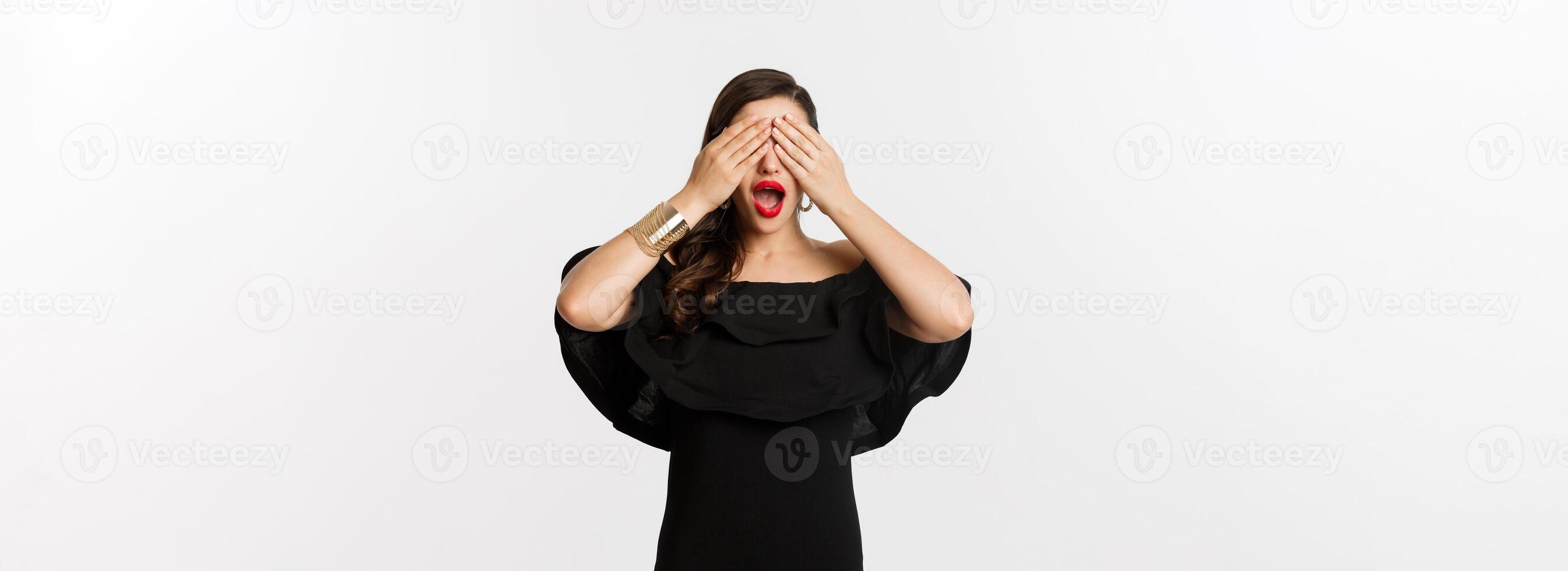 Fashion and beauty. Beautiful woman in black dress and red lipstick, covering eyes and smiling, waiting for surprise, standing over white background photo