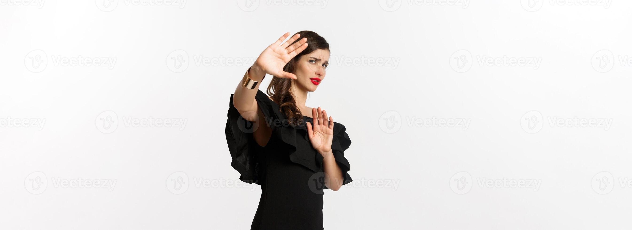 Fashion and beauty. Reluctant and worried woman asking to stay away, showing stop gesture and looking scared, standing in black dress over white background photo