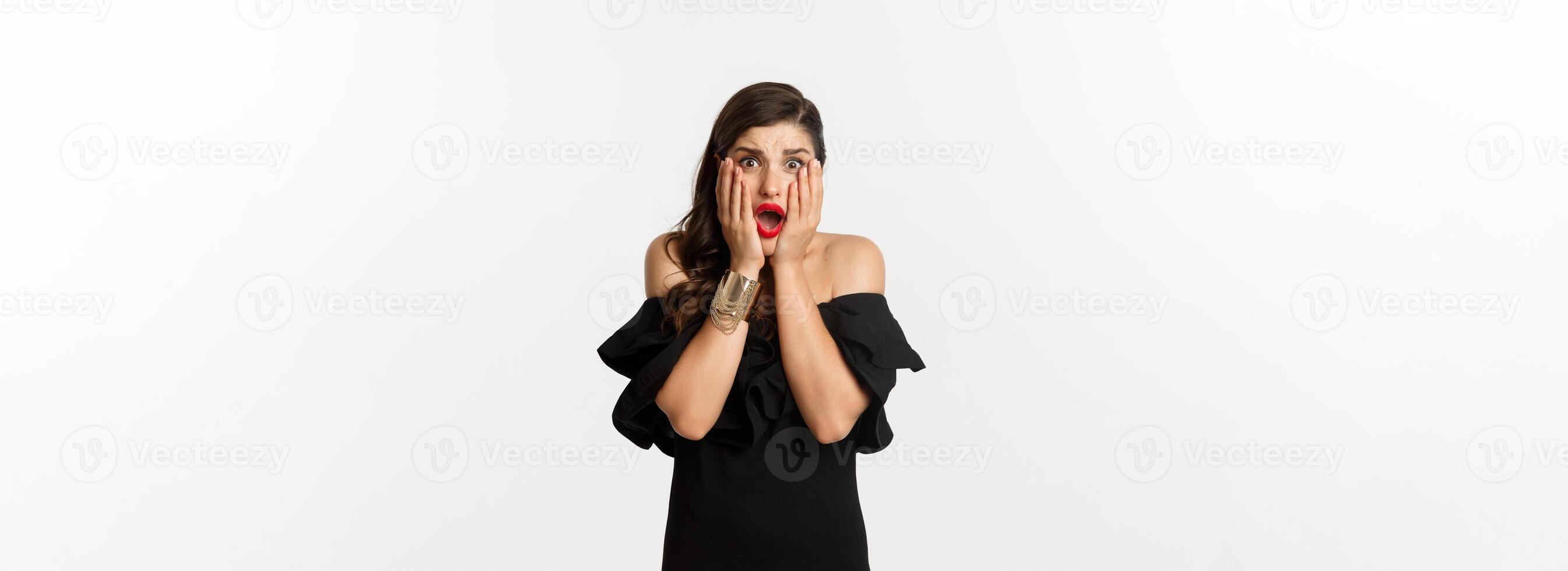 Fashion and beauty. Shocked and worried woman in black dress looking scared, standing anxious over white background photo