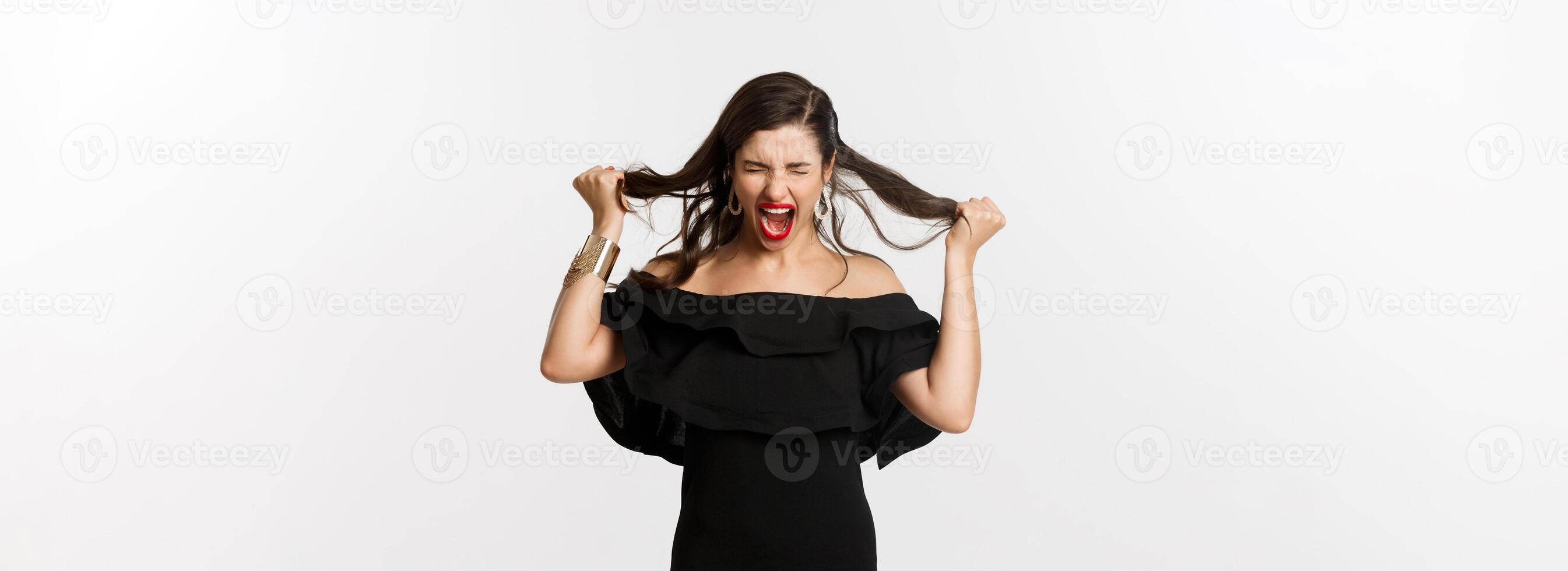 Fashion and beauty. Young woman in black dress screaming and rip hair on head, screaming mad, standing angry and outraged over white background photo