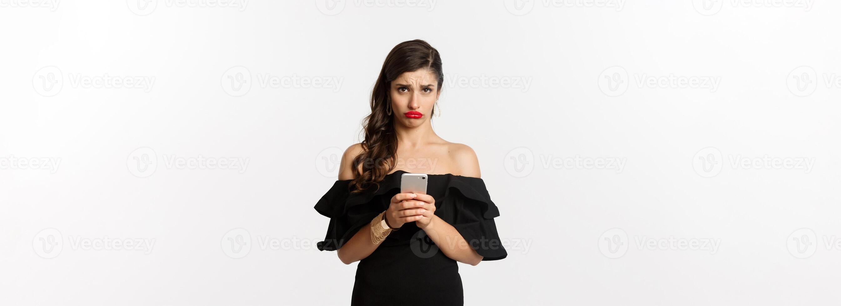 Sad and gloomy woman in black dress, sulking upset, using mobile phone and feeling disappointed, standing over white background photo