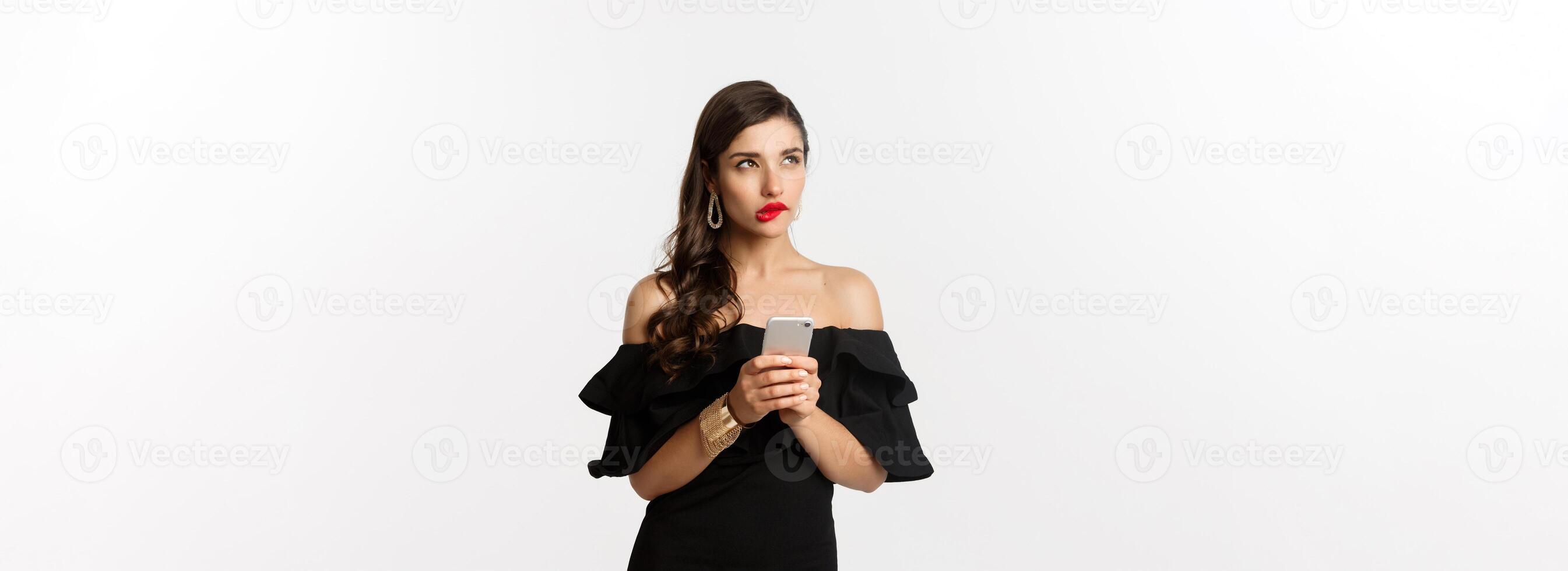 Thoughtful young woman using smartphone and thinking, looking up and pondering, standing over white background photo