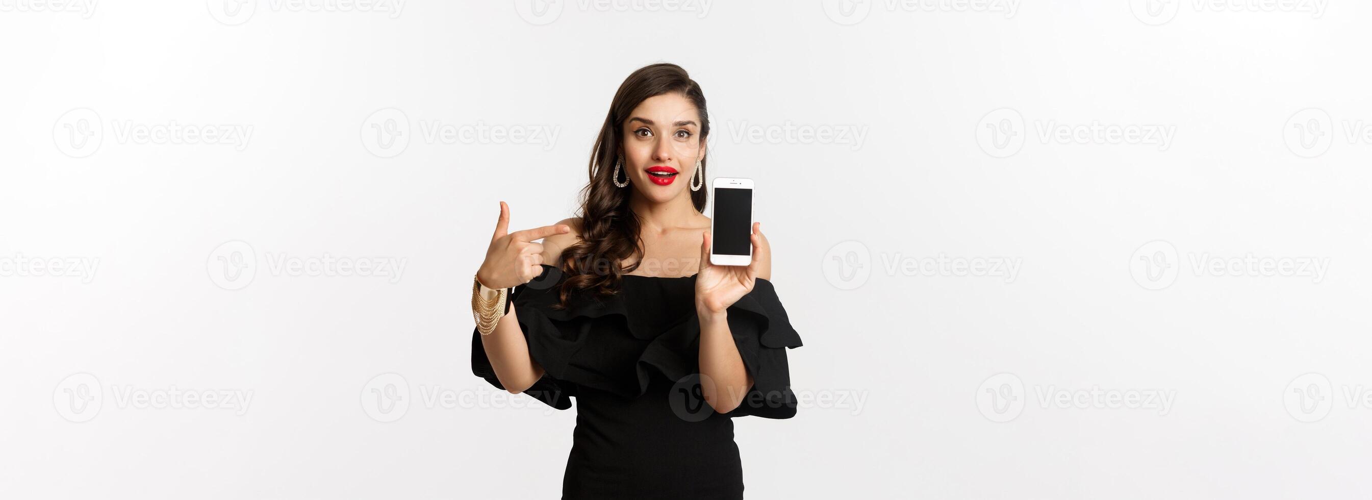 Online shopping concept. Fashionable woman in black dress pointing finger at smartphone screen, showing application, standing over white background photo