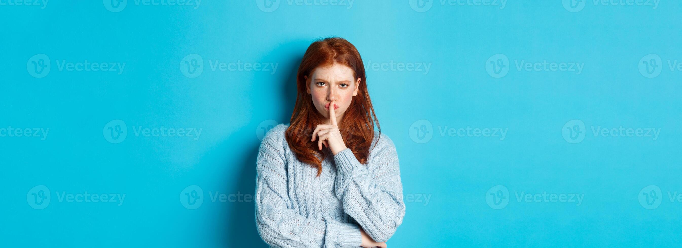 Angry redhead girl hushing at you, show taboo gesture, forbid to speak, standing over blue background in sweater photo