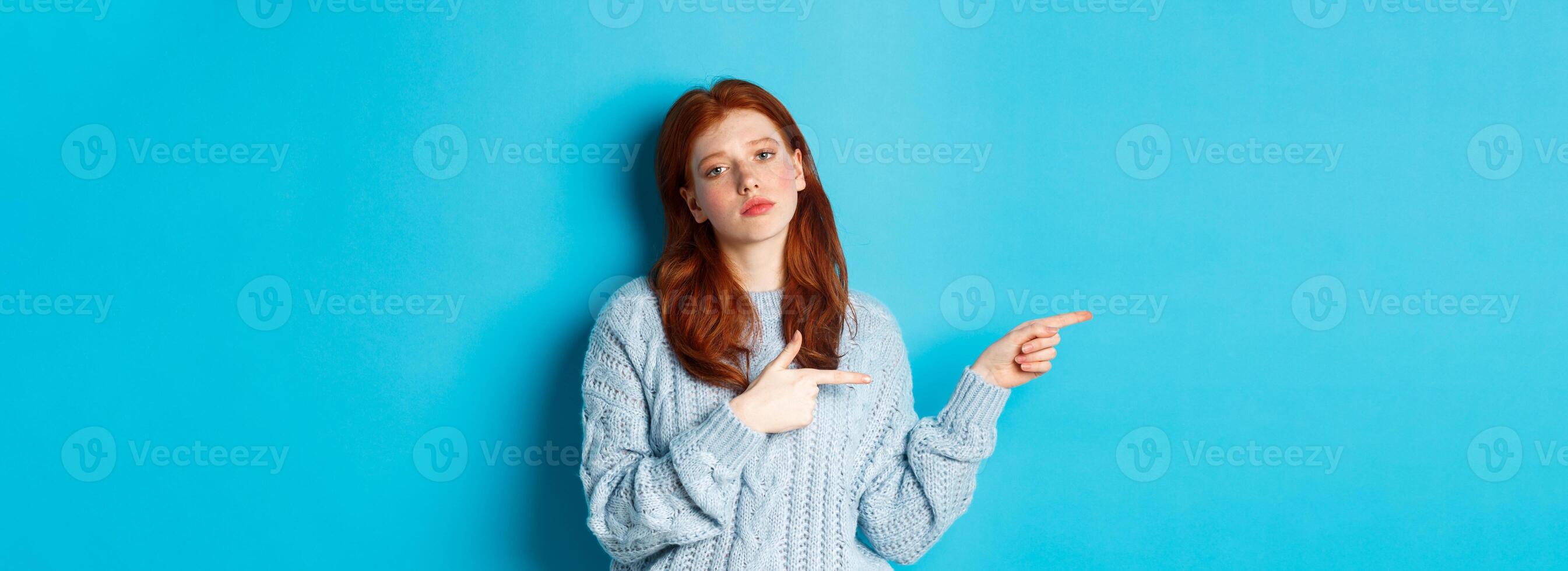 Moody teenage girl with red hair, pointing fingers left at logo, staring bothered and bored, standing over blue background photo