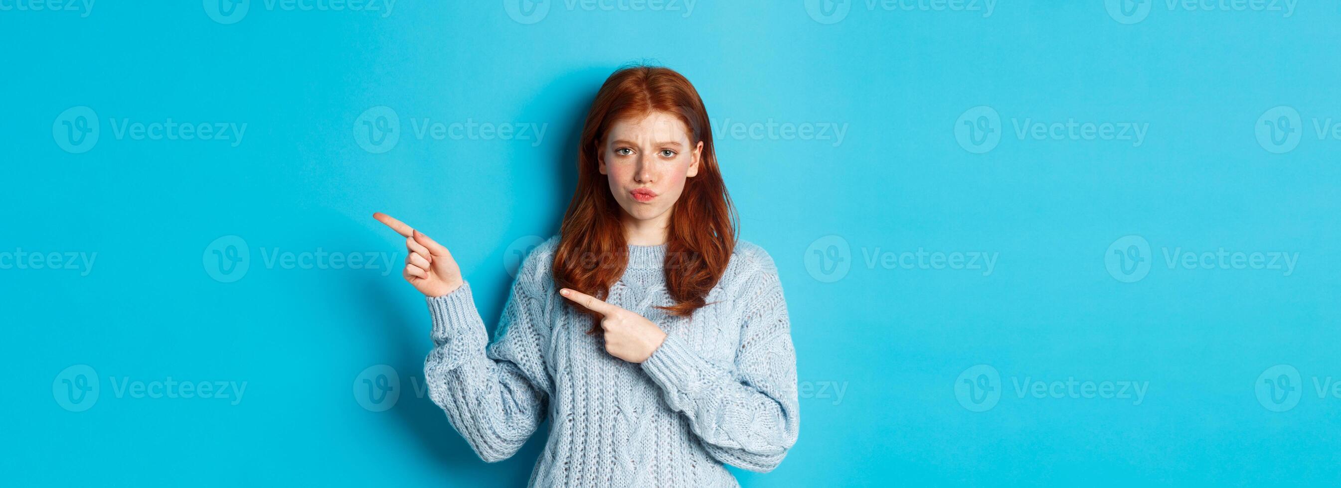 Winter holidays and people concept. Complicated redhead girl staring indecisive, pointing fingers right at logo and frowning, standing perplexed against blue background photo