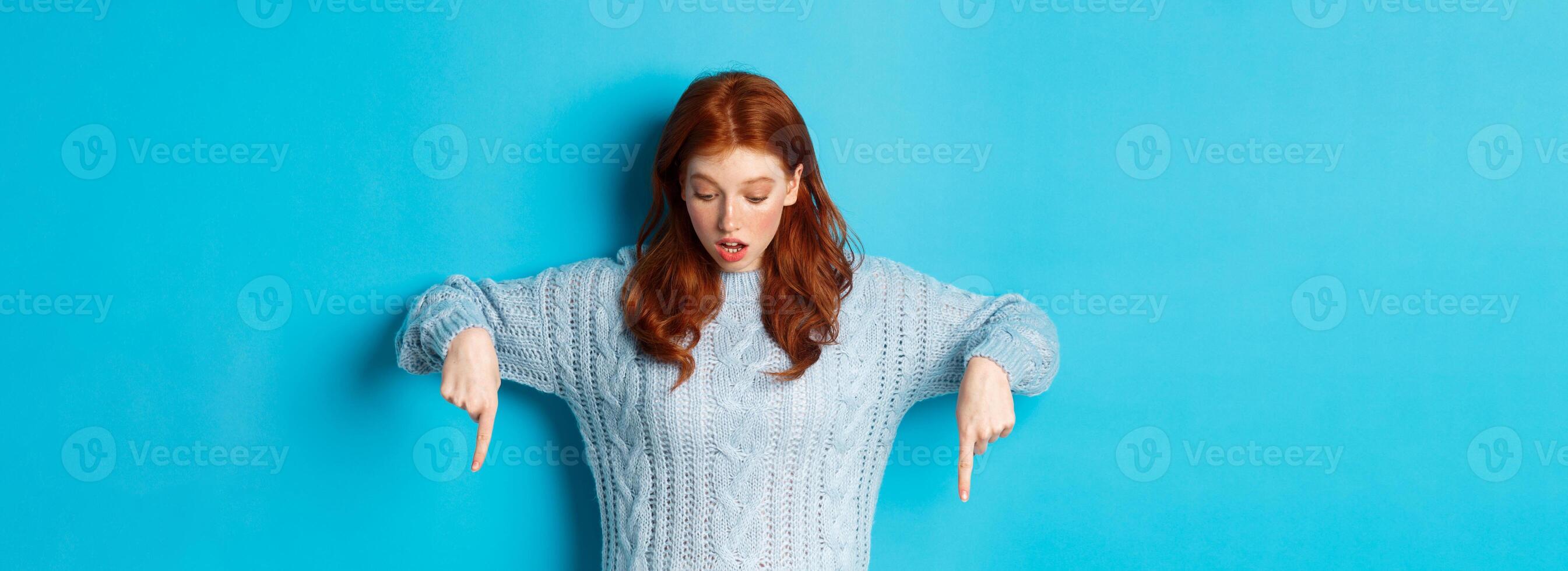 Winter holidays and people concept. Impressed redhead girl in sweater, looking and pointing down with amazement, standing against blue background photo