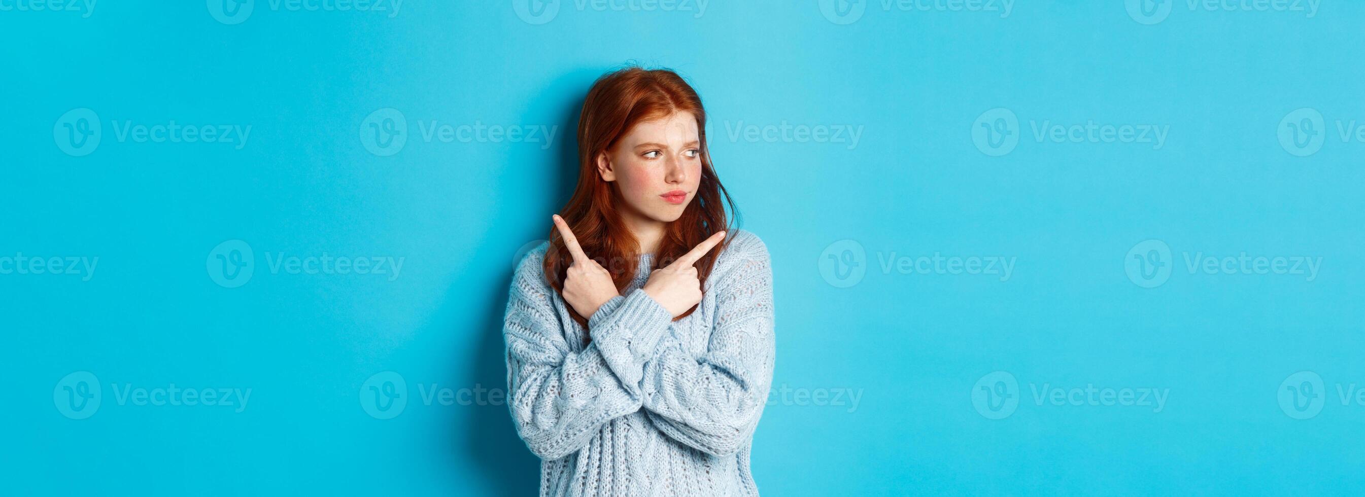 Indecisive redhead teenage girl making decision, pointing fingers sideways and looking left doubtful, standing in sweater against blue background photo