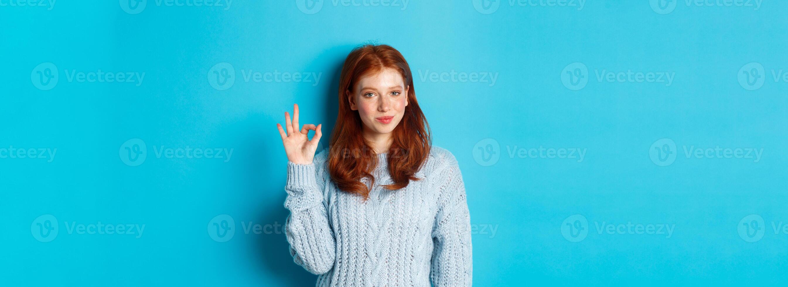 Confident redhead girl assuring you, showing okay sign and smiling, saying yes, approve and agree, standing over blue background photo