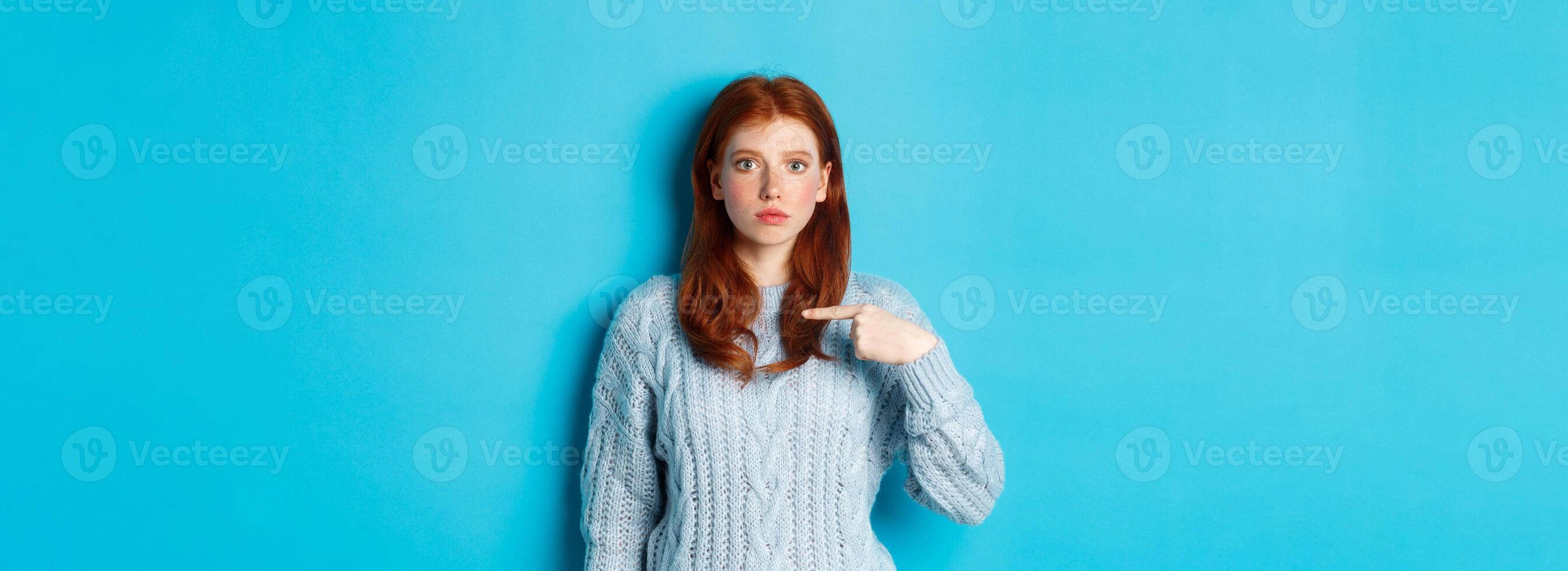 Nervous and confused redhead girl pointing at herself, standing in sweater against blue background photo