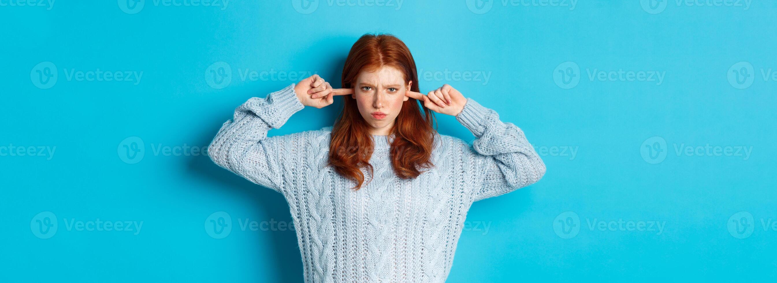 Teenage redhead girl unwilling to listen, shut ears and frowning angry, staring at camera offended, sulking against blue background photo