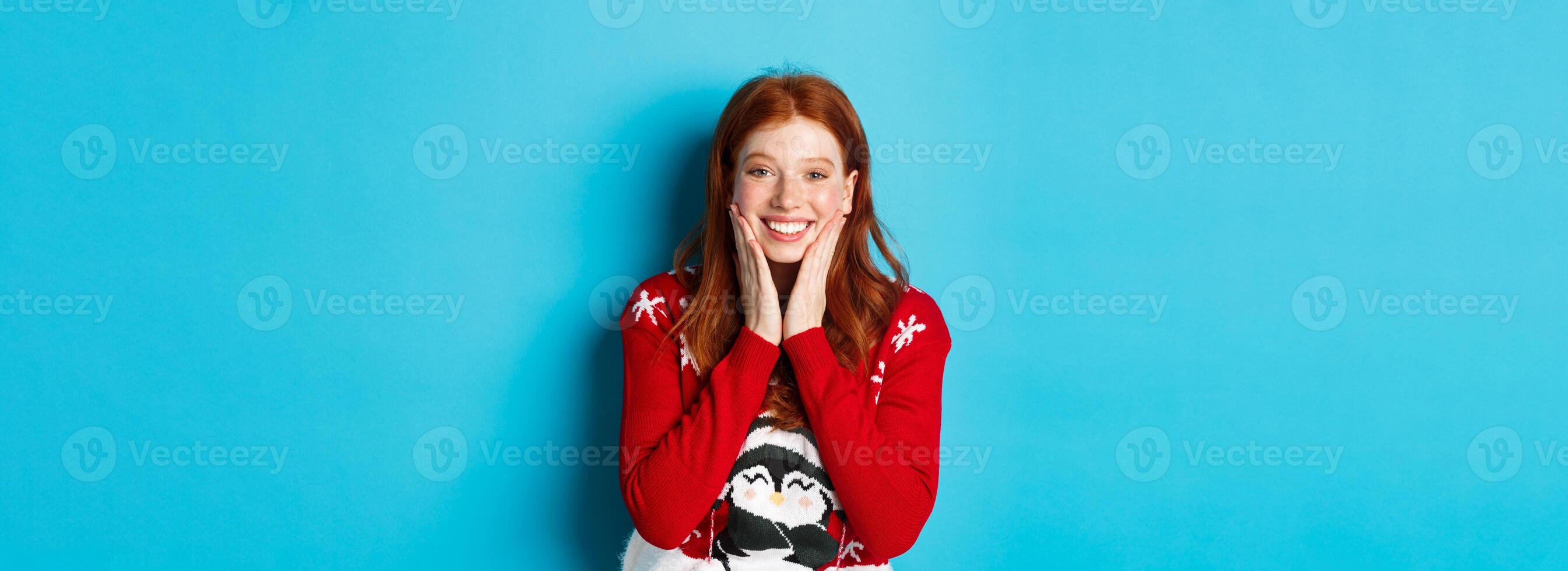 Winter holidays and Christmas Eve concept. Adorable redhead girl blushing and touching cheeks, staring grateful and happy at camera, standing in xmas sweater against blue background photo