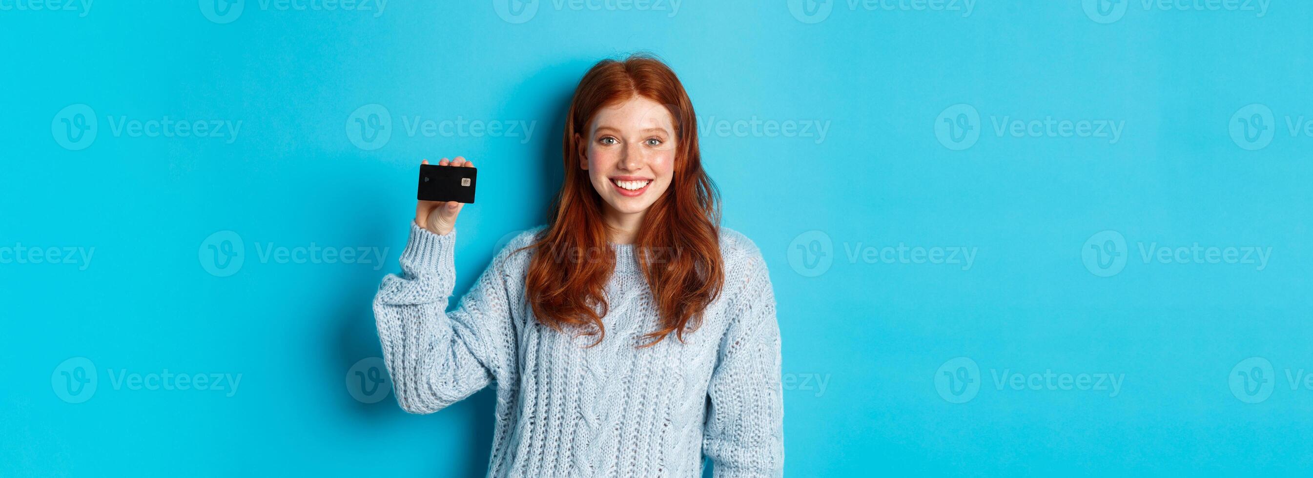 Cute redhead girl in sweater showing credit card, smiling at camera, standing over blue background photo