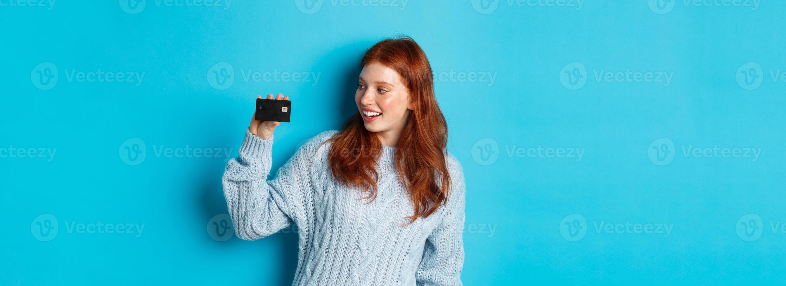 Cute redhead girl in sweater showing credit card, smiling at camera, standing over blue background photo