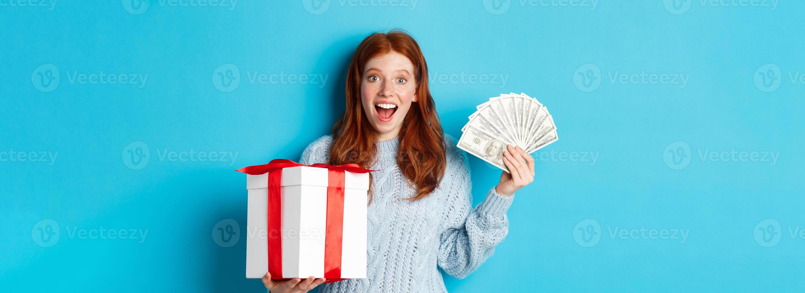 Christmas and shopping concept. Happy redhead woman holding money and big xmas present, showing dollars and gift, smiling pleased, standing over blue background photo