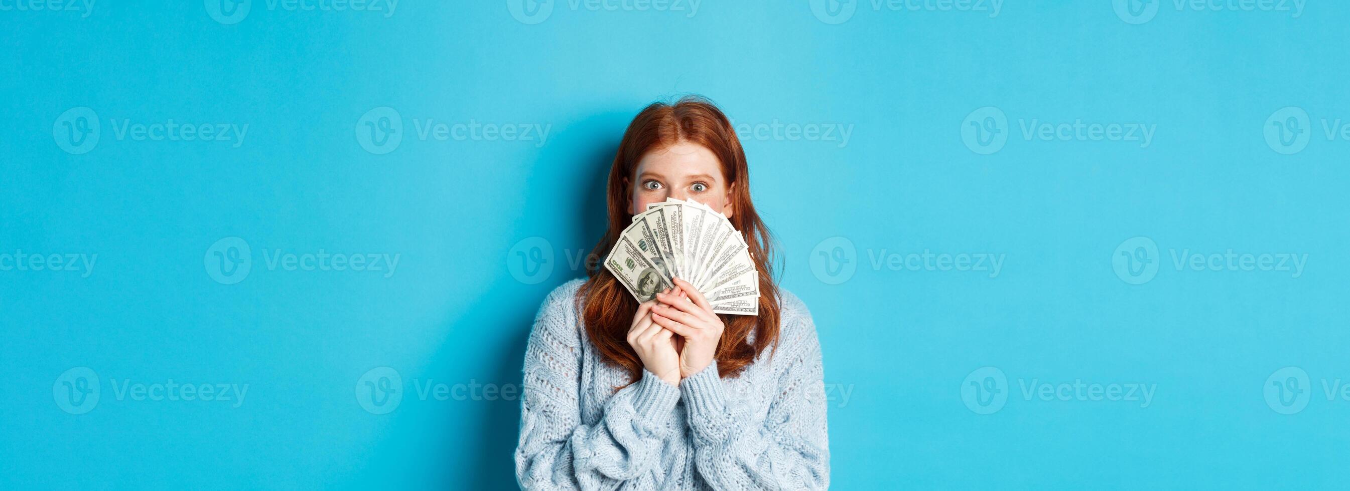 Excited redhead woman covering face with money, holding dollars and staring at camera happy, standing over blue background photo