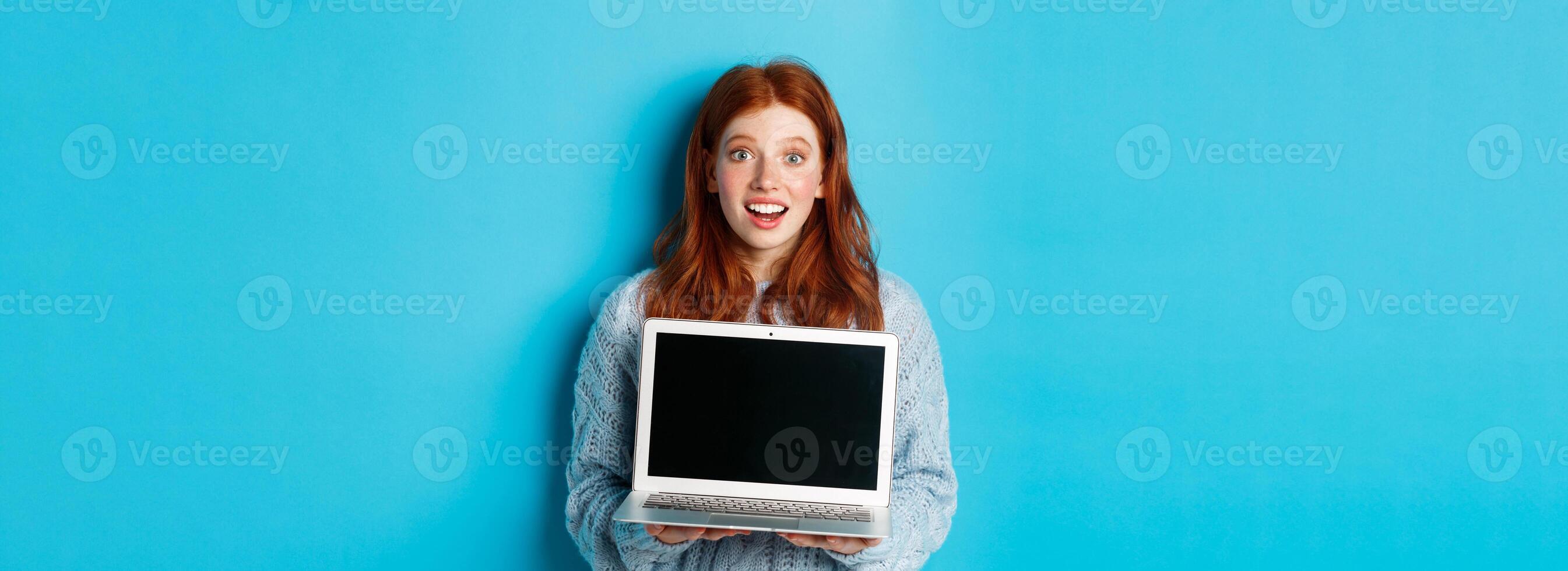 Excited redhead female freelancer showing laptop screen, staring at camera amazed, standing with computer against blue background photo