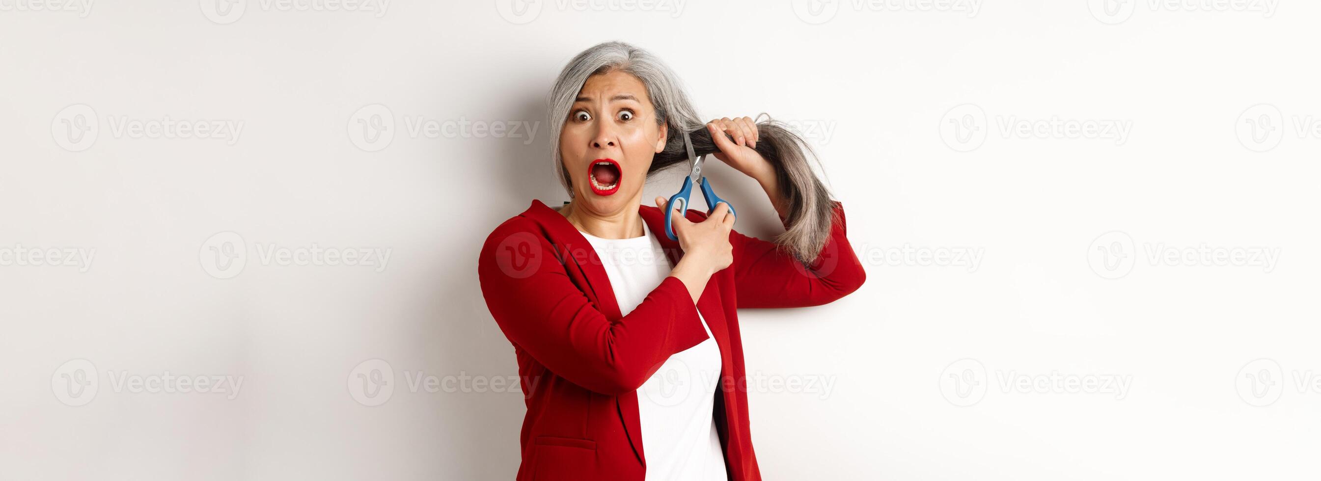 Scared asian senior woman cutting hair with scissors and screaming in panic, standing over white background photo