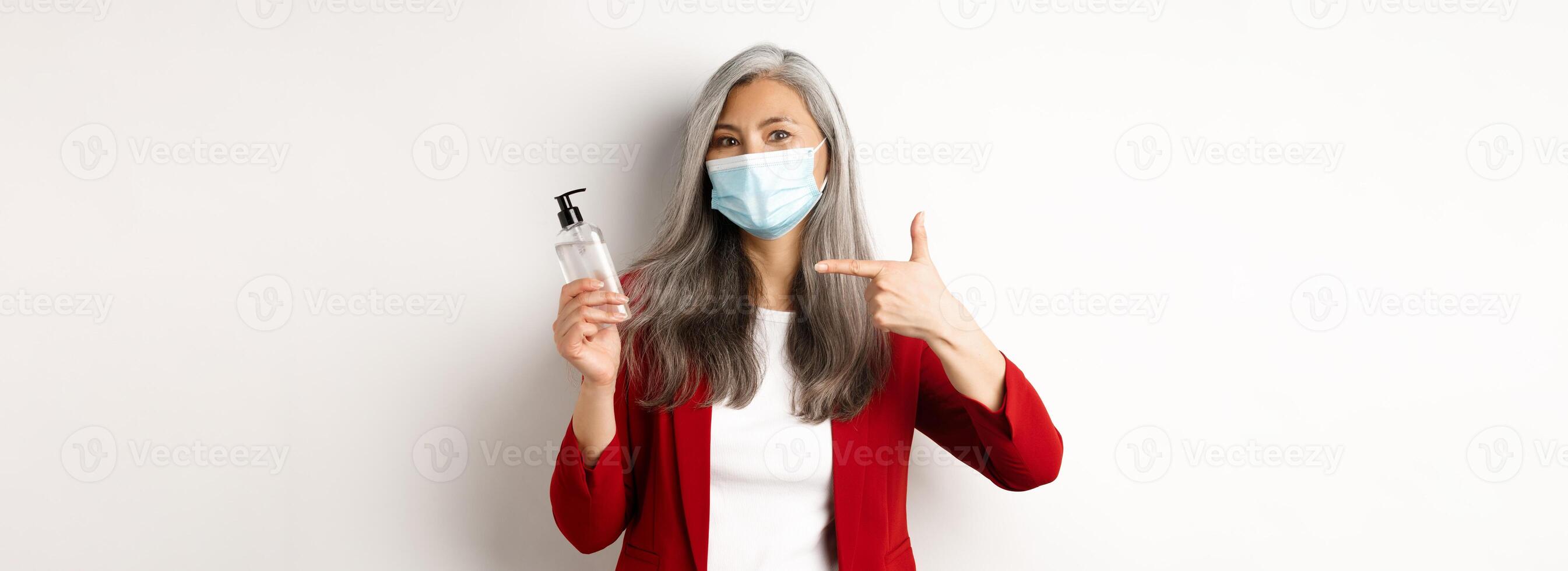 Covid, pandemic and business concept. Asian female manager in face mask, pointing finger at hand sanitizer, recommending antiseptic, white background photo