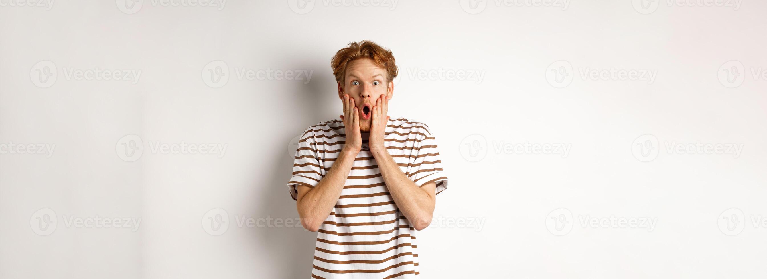 Impressed young man with ginger hair, gasping and staring shocked at camera, express complete disbelief and amazement, white background photo