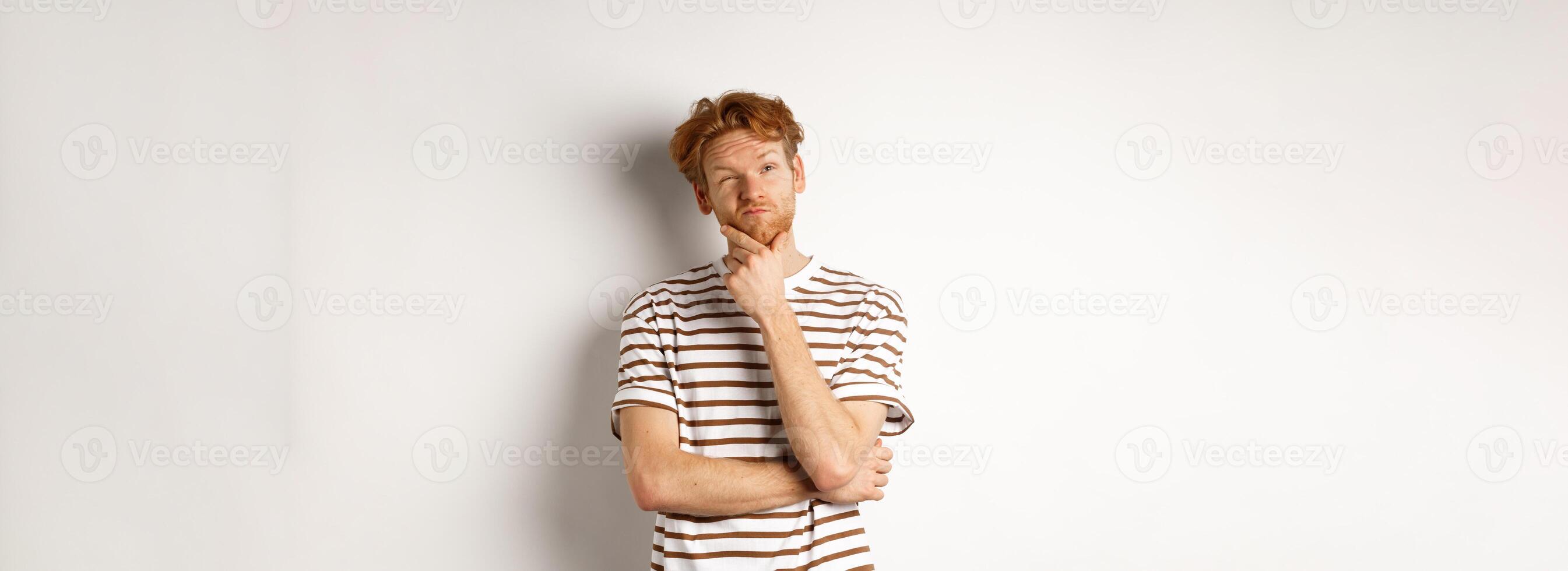 Portrait of thoughtful man with red hair thinking, looking at upper left corner and making choice, standing over white background photo