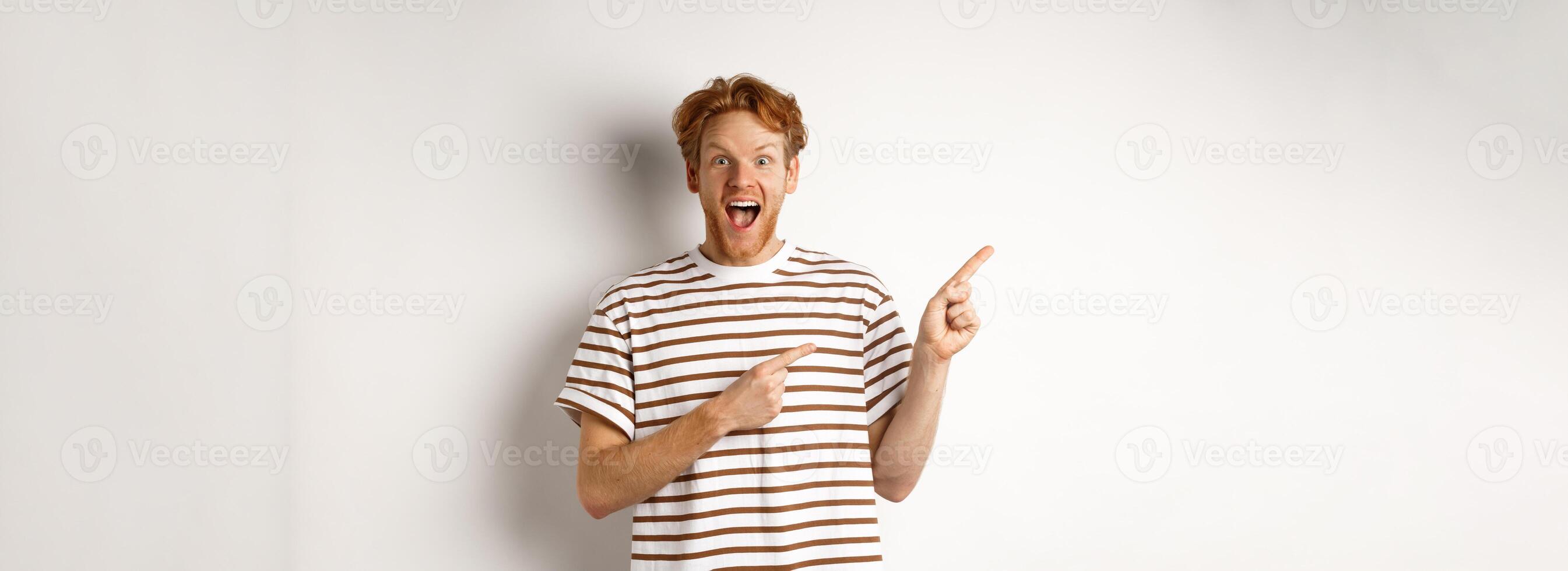 Cheerful man with red hair checking out promo offfer, pointing left at logo and smiling at camera, standing over white background photo