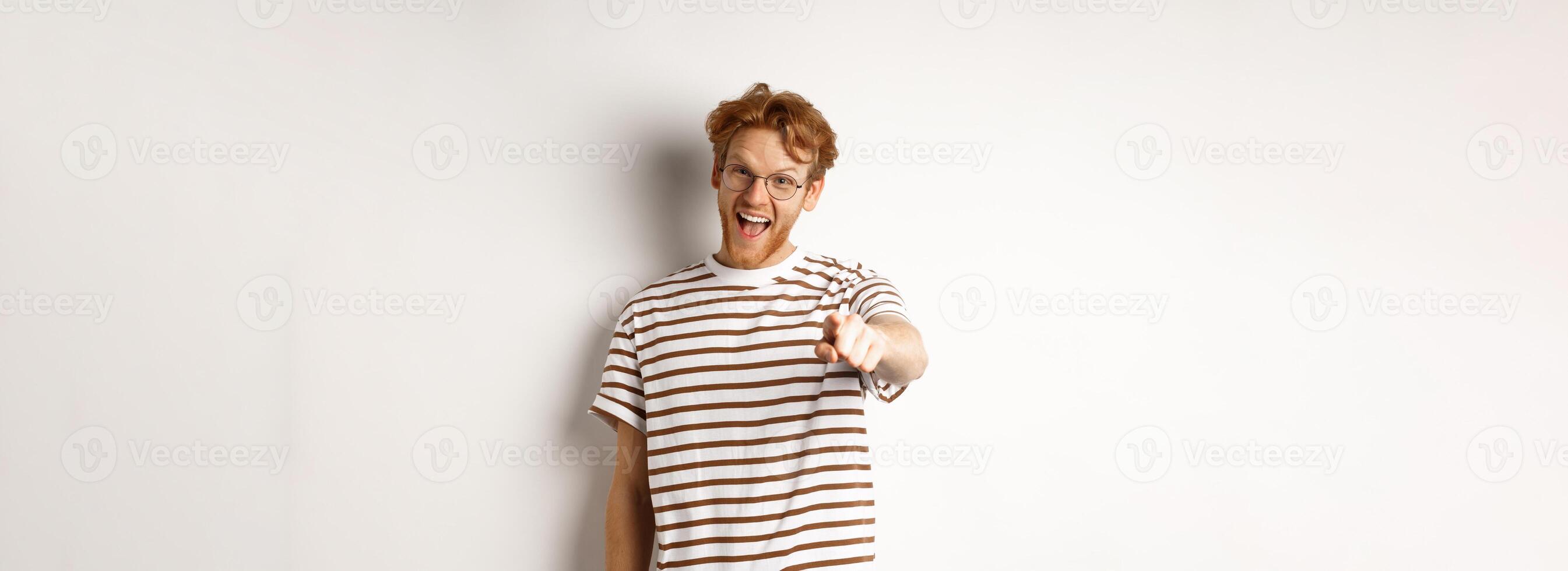 Image of funny redhead guy in glasses laughing at you, pointing finger camera and making fun of someone, standing over white background photo
