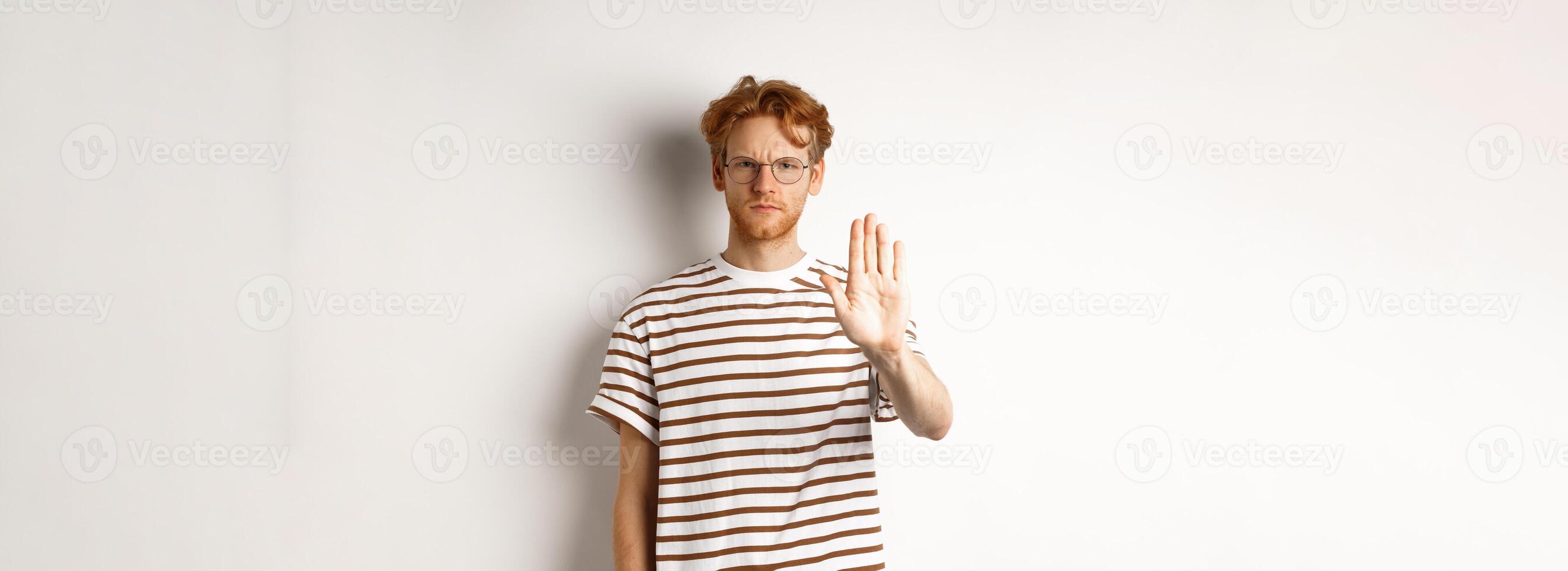 Serious young man with ginger hair frowning, showing stop gesture, refuse and prohibit something bad, standing displeased over white background photo