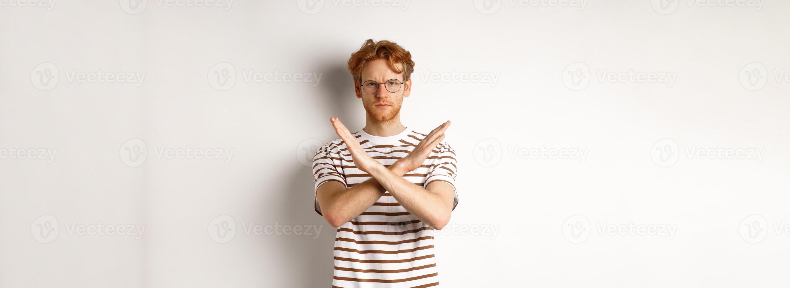 Serious young man with ginger hair frowning, showing stop gesture, making cross to prohibit something bad, disagree with you, standing over white background photo