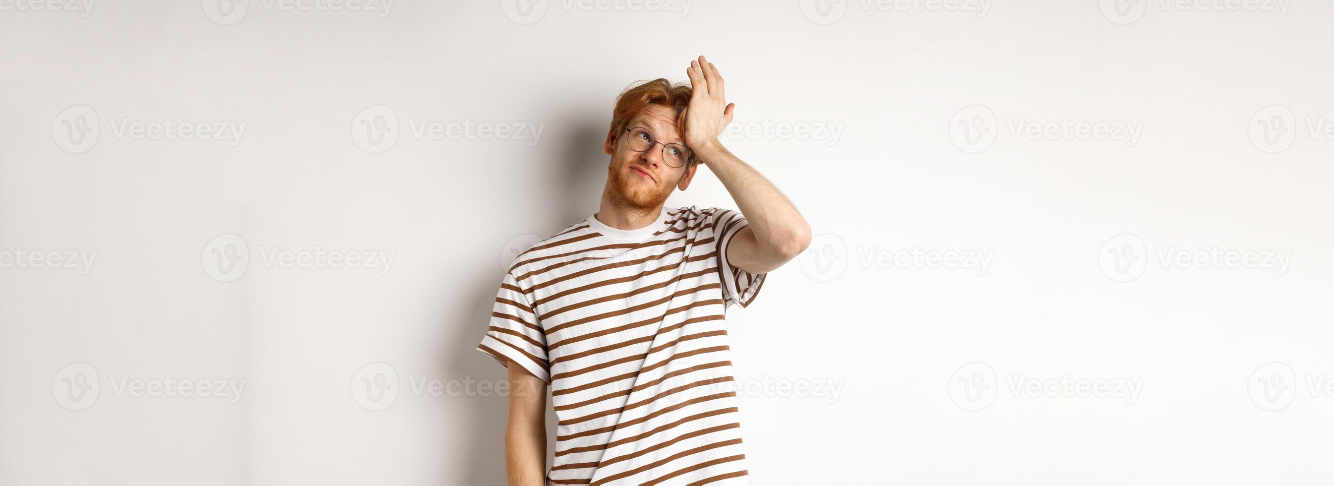 Tired young man with red hair and glasses, roll eyes and making facepalm bothered, standing over white background photo